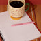 cream short ceramic mug with pink cursive 'in my growing season' with green handle and pink and yellow pedestal on a notebook and filled with coffee