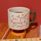 cream short ceramic mug with pink cursive 'in my growing season' with green handle and pink and yellow pedestal on a table filled with coffee