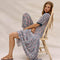 model wearing pink and blue floral midi dress with tiered skirt, puffed short sleeves and blue trim on the neckline and sleeves while sitting in a chair
