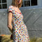side view of model wearing white cotton mini dress with red lettuce trim, bow on the front bust, and retro primary color floral print