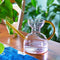light pink abstract shaped watering can with amber colored spout and handle on a table
