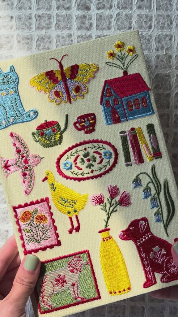 model showcasing cream embroidered journal wth various folk items - homes, butterflies, stamps, dog, cat, books